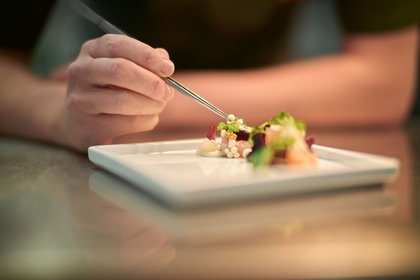 Eine Person dekoriert ein kunstvoll angerichtetes Gourmetgericht auf einem weißen Teller im Restaurant in Stolberg im Harz