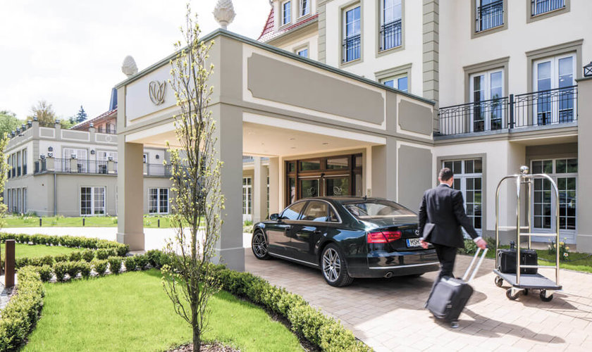 Ein eleganter Hoteleingang, an dem ein Gast mit Koffer eintrifft, während ein Auto vor dem Eingang geparkt ist