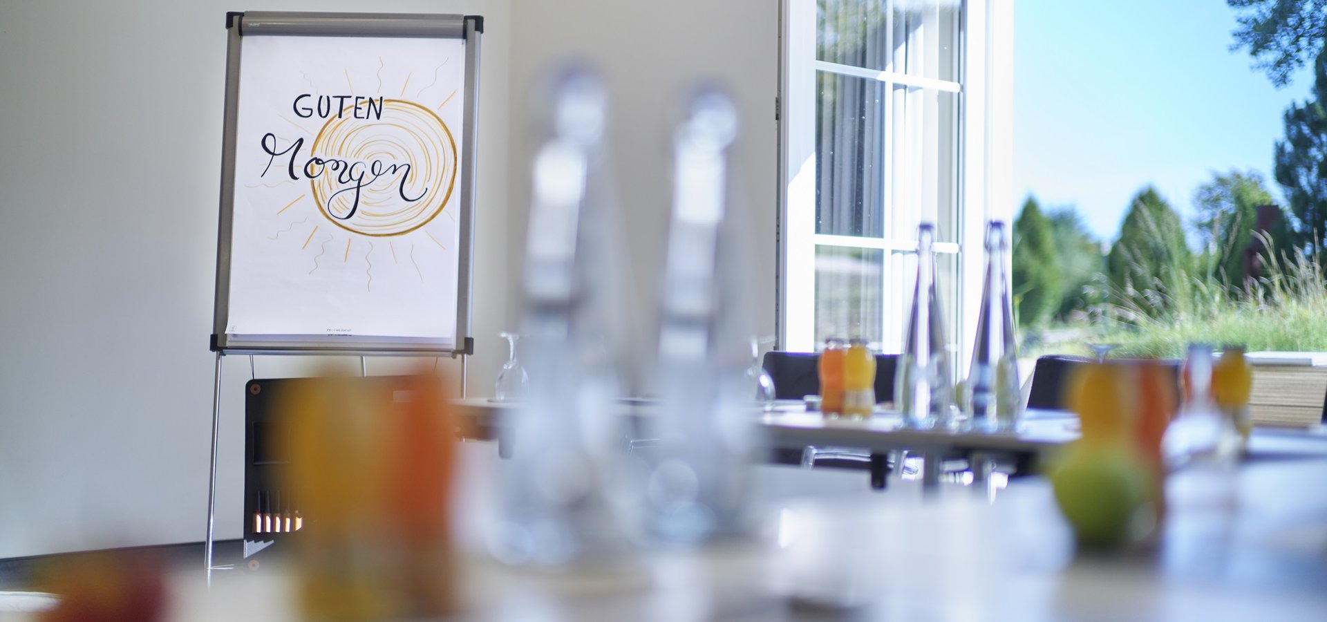 Flipchart mit der Aufschrift "Guten Morgen" in einem hellen Tagungsraum mit Getränken auf dem Tisch und Blick ins Grüne