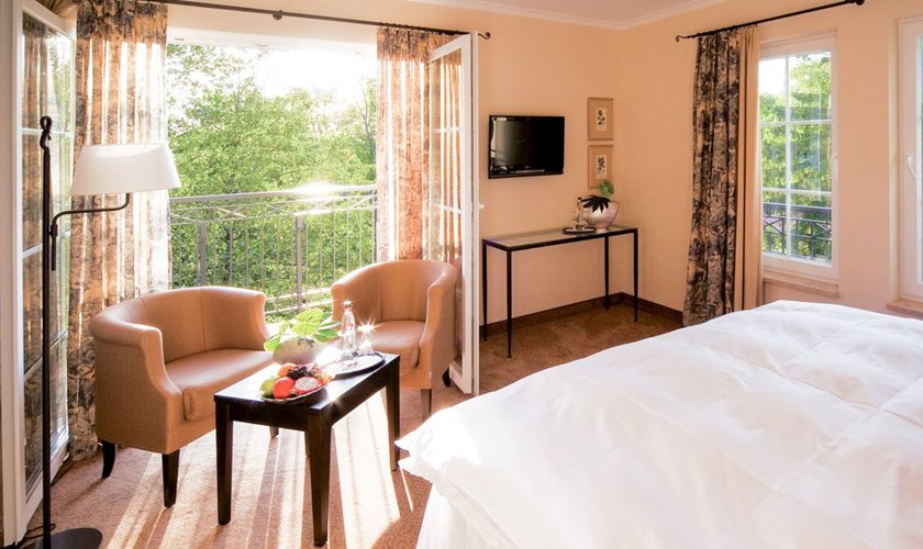 Ein lichtdurchflutetes Zimmer mit offenem Balkon, Sonnenstrahlen kommen rein, Blick in die Natur und einem Doppelbett und Sessel und Tisch.
