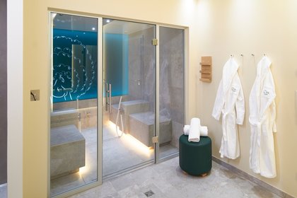 Moderner Wellnessbereich mit Glasdampfbad, hellen Fliesen und zwei weißen Bademänteln an der Wand