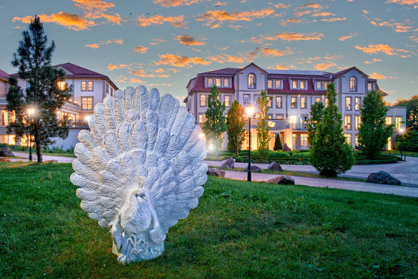 Eine weiße Pfauenskulptur steht im Vordergrund eines gepflegten Gartens bei Sonnenuntergang, mit einem eleganten Gebäude im Hintergrund.