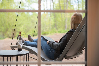 Eine Frau liegt in einer Hängeliege vor der Landresidenz im Schindelbruch, schaut in die Natur und genießt ihren Wein.