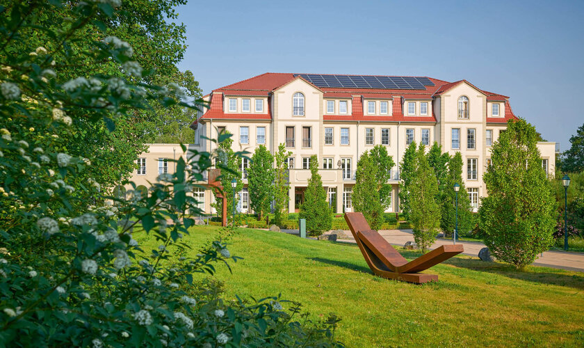 Ein elegantes Gebäude mit roten Dächern und Solarpanelen, umgeben von einer grünen Gartenlandschaft mit modernen Holzliegen