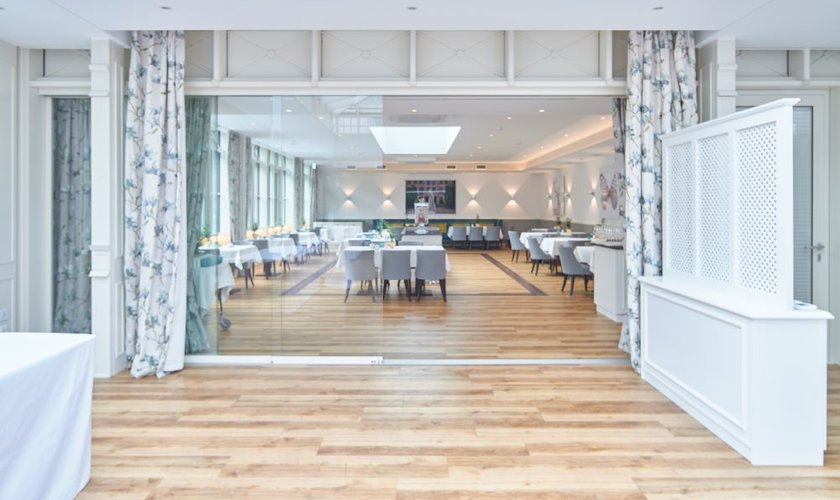 Lichtdurchfluteter Konferenzraum im Wintergarten, mit weißer Einrichtung und hellbraunem Parket. Dieser Raum kann wird auch als Restaurant Kleiner Waldteufel genutzt.
