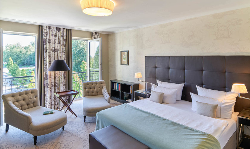 Ein elegantes Hotelzimmer mit einem großen, gemütlichen Bett, zwei Sesseln und einem Balkon. Ein elegantes Hotelzimmer mit einem großen, gemütlichen Bett, zwei Sesseln und einem Balkon mit Blick ins Grüne.